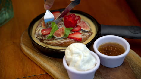 Slow-motion-of-a-female-cutting-through-a-cinnamon-roll-topped-with-sliced-figs-and-strawberries-in-an-iron-skillet-accompanied-with-whipped-cream-and-sauce