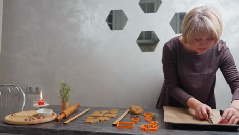 woman placing cutout shaped dough pieces onto baking tray, preparing for baking in oven, surrounded by kitchen tools, cookie cutters, and aromatic ambiance with candle and rosemary in background