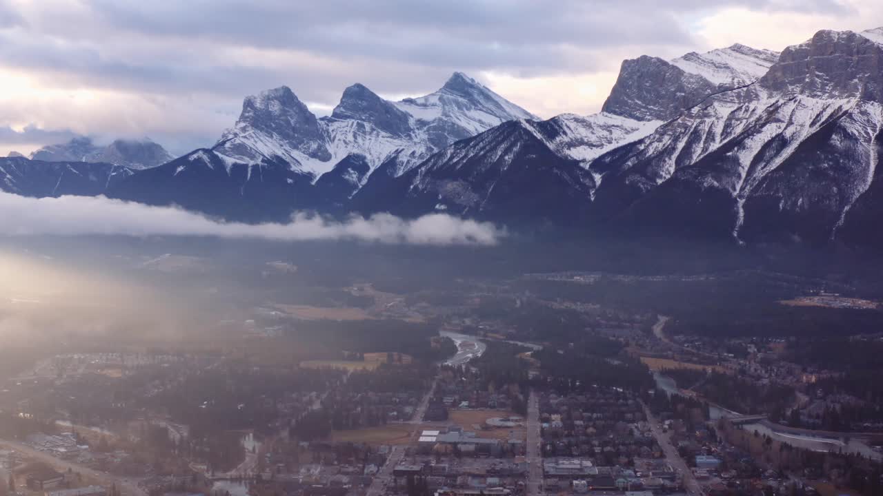 Slow Dramatic Left Pan Over Town Of Canmore In A Valley In Alberta, Canada,  As The Sun Rises Over Low Clouds, With Three Sisters Mountain Range  Towering In The Background Free Stock