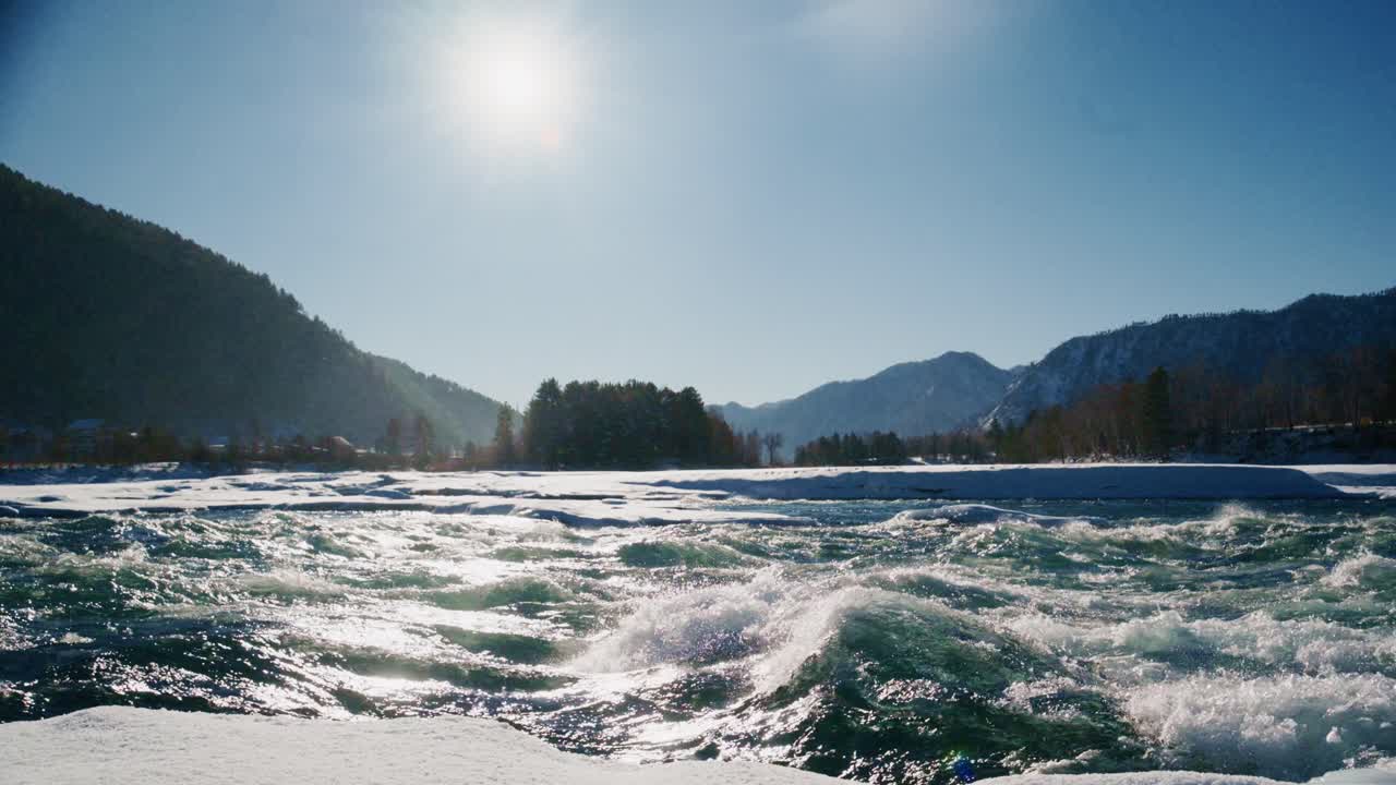 川沿いの素晴らしい冬の朝の風景。氷の張った川岸、流れる水、そして波に反射する鮮やかな太陽光が目に飛び込んできます。雄大な山々が穏やかな風景を縁取ります。