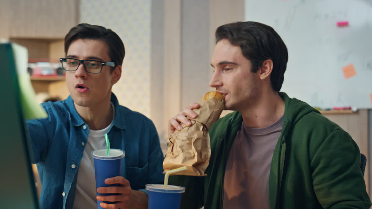 샌드위치를 먹는 사무실 직원들이 근접한 모니터를 보고 있습니다. 활발한 노동자들이 토론하고 있습니다.