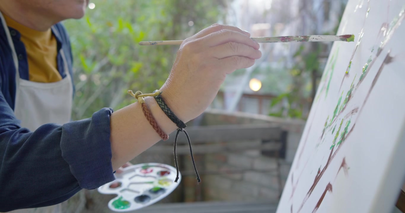 汚れたエプロンをつけた高齢のアジア人男性が、ベランダでキャンバスに葉を描き、絵の具に筆を浸している。