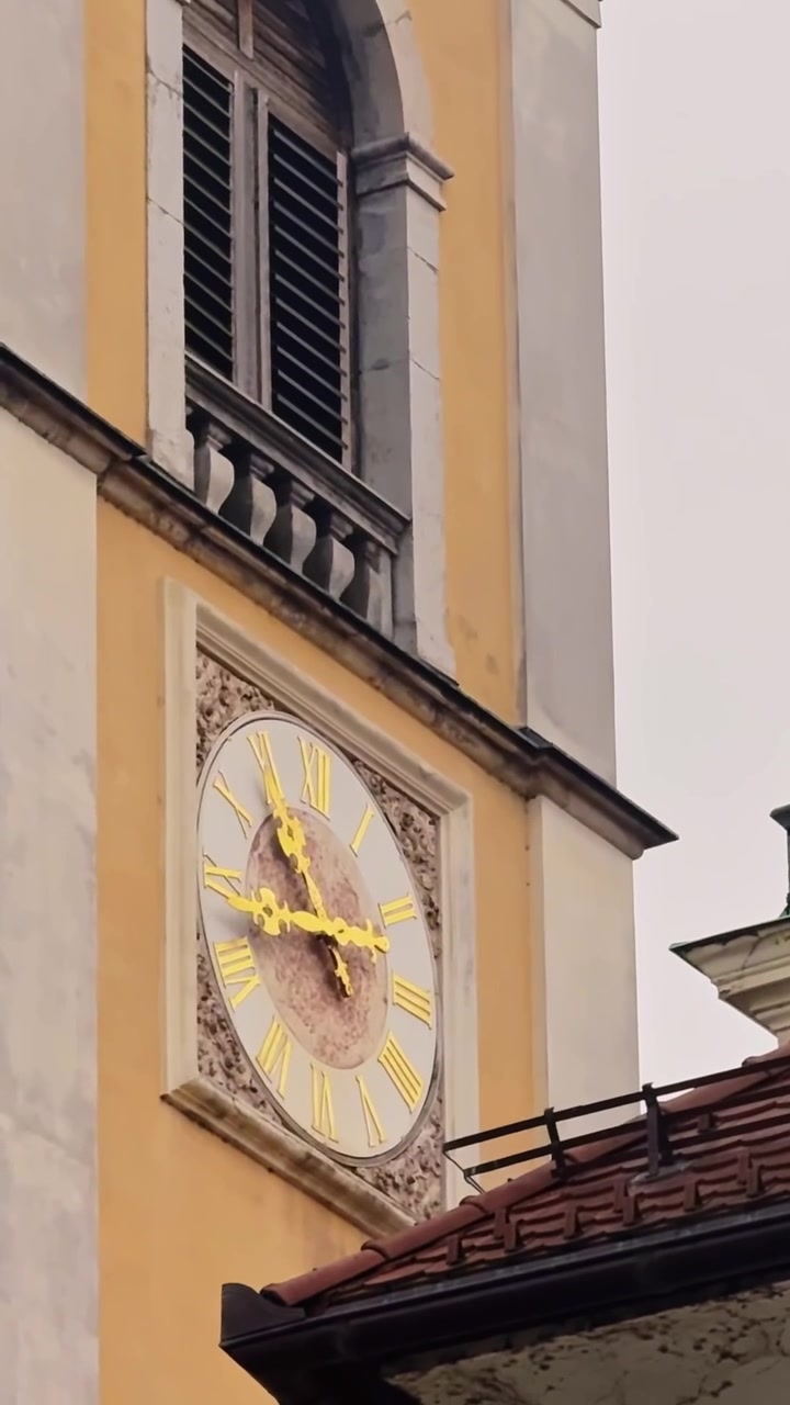 Primo piano verticale del campanile della Cattedrale di Lubiana, che mostra un orologio con numeri romani e dettagli dorati, un'apertura ad arco con persiane e una facciata barocca gialla