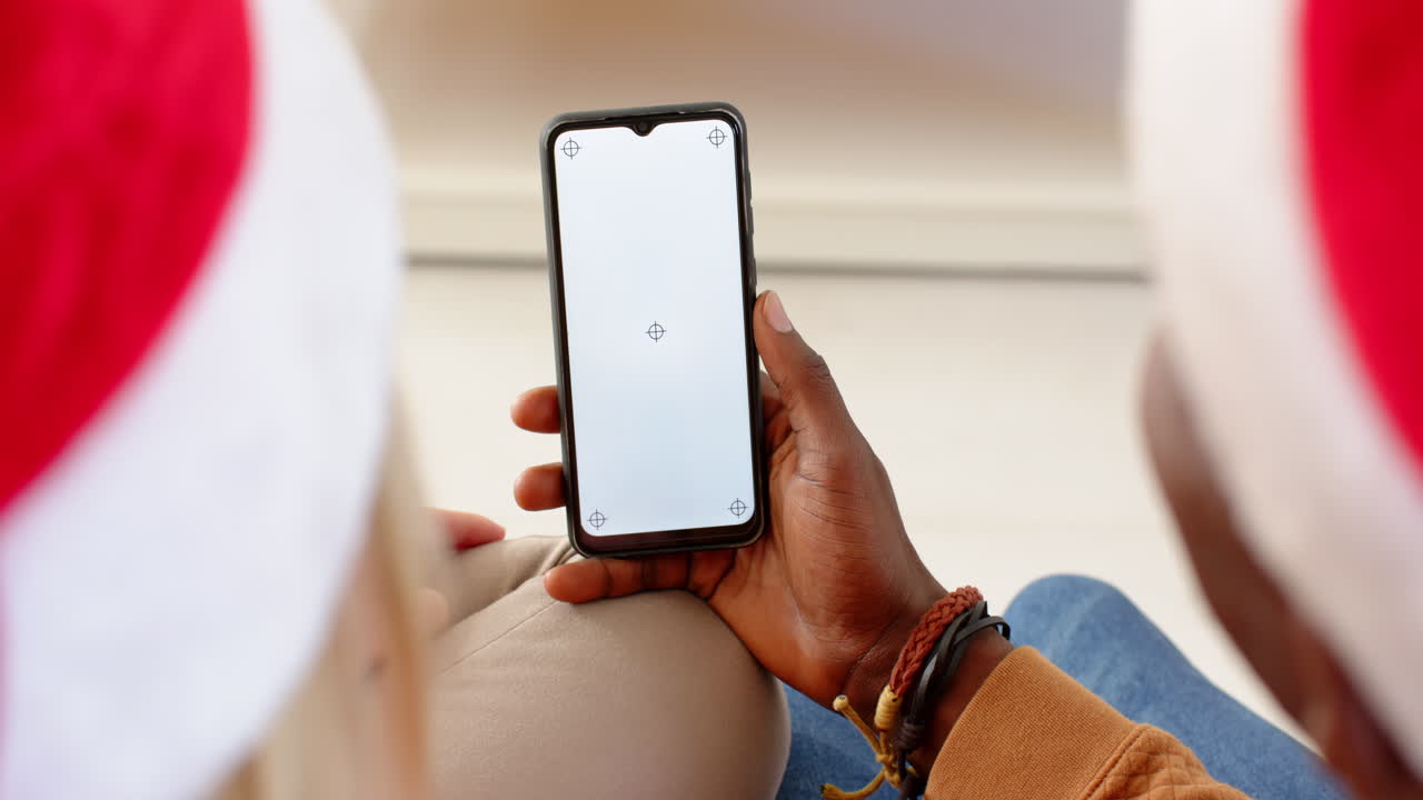 Coppia multietnica che indossa cappelli di Babbo Natale e usa lo smartphone insieme durante la celebrazione del Natale, con spazio per il testo