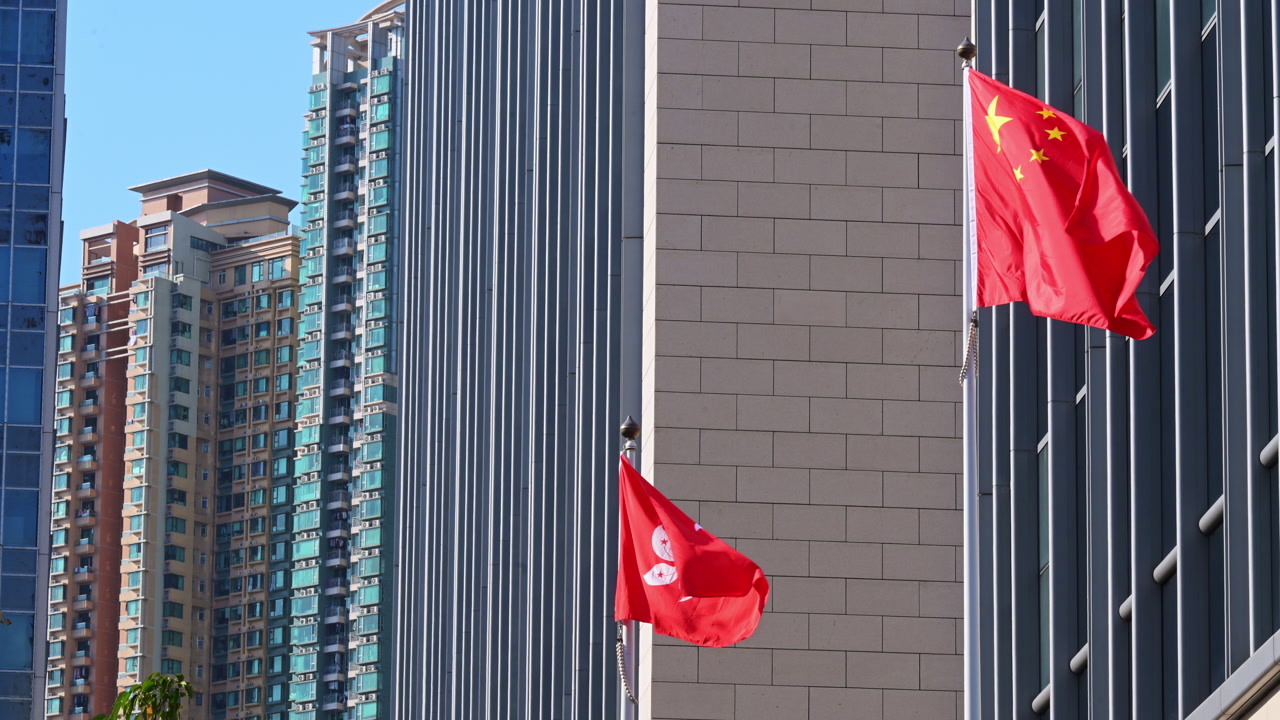 People's Republic of China and Hong Kong SAR flags wave against a backdrop of modern skyscrapers. Establishing shot of national identity and administrative sovereignty in a major Asian hub.