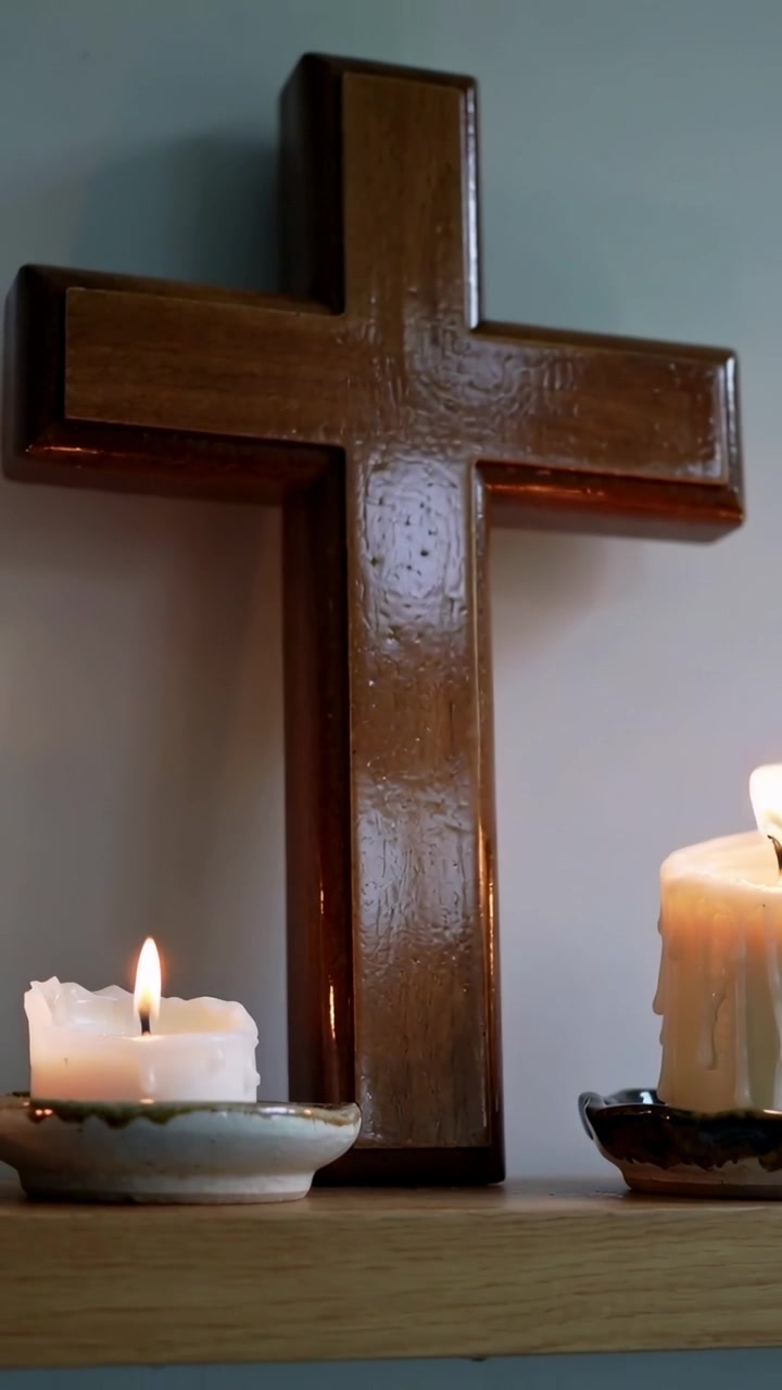 Video vertical: Cámara alejándose para revelar una cruz de madera en un estante, con dos velas encendidas parpadeando.