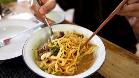 a person uses chopsticks and a spoon to mix beef curry noodles in a white bowl, under bright natural lighting at a restaurant table in bangkok