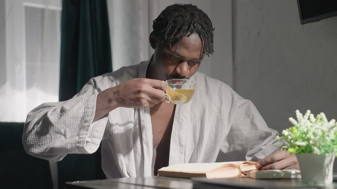 Man in bathrobe sitting at table, reading book, drinking tea, and relaxing. Indoor setting with natural light, green plants, and modern decor. Cozy, quiet, peaceful atmosphere perfect for mindfulness