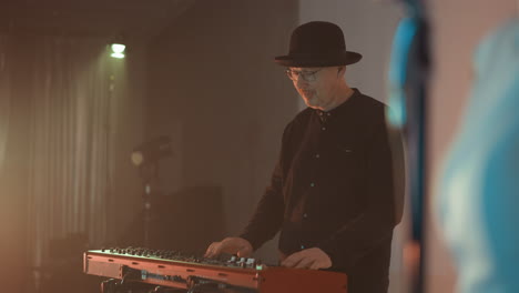 keyboardist in a black shirt, black hat, and glasses, playing a red sampler in a studio room with soft lighting. capturing the focused and immersive moment of music creation