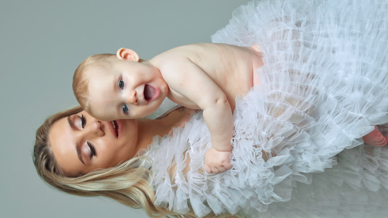 Adorable naked baby boy smiling sweetly sitting in mom's hands. Mother kisses her beloved son and presses to herself. Vertical screen.