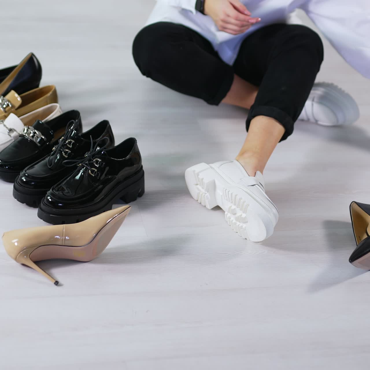 Unrecognizable lady in white shirt and black jeans sits on the floor and picks footwear. Various kinds of shoes are on the floor around female. White backdrop