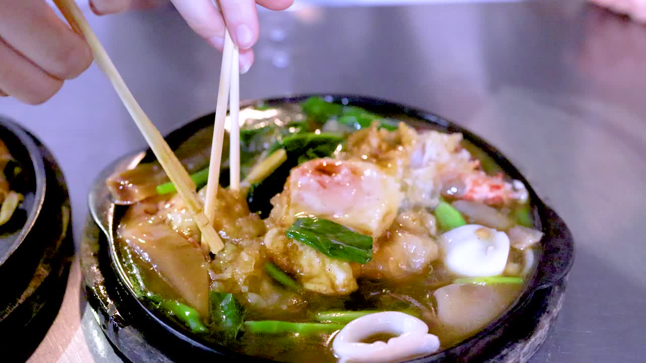 Hand uses chopsticks to pick up seafood pad thai noodles from a sizzling hot plate