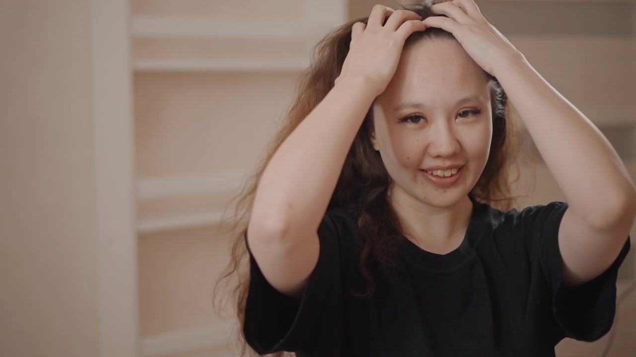 Potret Dalam Ruangan: Wanita Muda Tersenyum Menata Rambut Panjang Bergelombang dengan Tangan dan Sikat dalam Cahaya Rumah yang Hangat