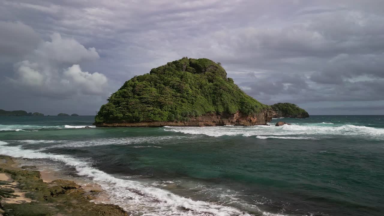 Un filmato aereo mostra un piccolo isolotto tropicale nell'oceano aperto con onde che si muovono, una spiaggia rocciosa, una ricca vegetazione e nuvole mutevoli che modellano un paesaggio costiero incontaminato a Giava Orientale.