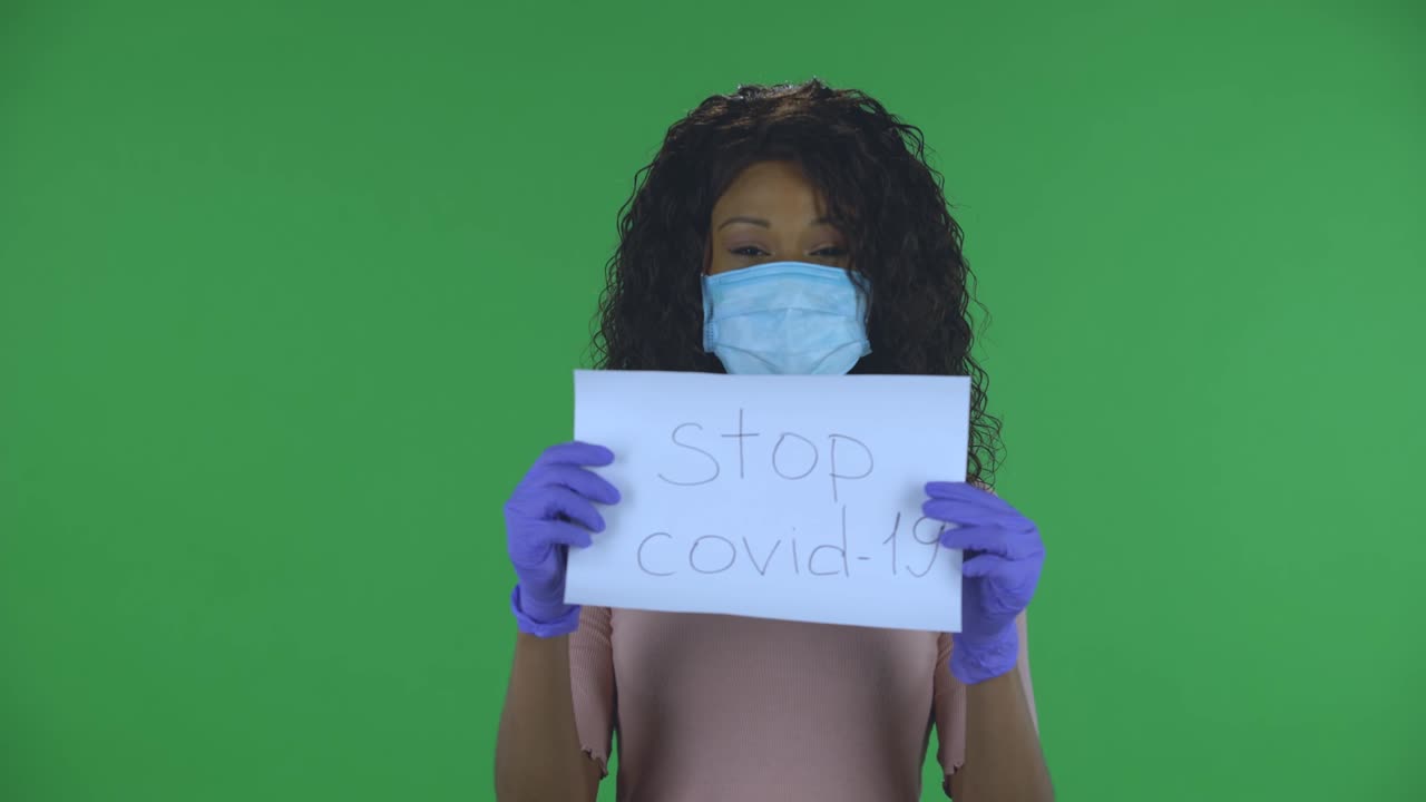 Portrait of beautiful african american young woman in medical mask is looking at camera and holding a poster Stop Kovid-19. Burning brunette with wavy hair in jeans and a beige blouse on a green screen in the studio. Health Protection Corona Virus Concept