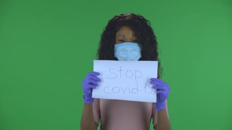 portrait of beautiful african american young woman in medical mask is looking at camera and holding a poster stop kovid-19. burning brunette with wavy hair in jeans and a beige blouse on a green screen in the studio. health protection corona virus concept