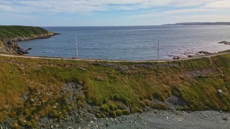 A drone glides over Ferryland&#039;s grassy headland, capturing the gentle slope meeting calm blue waters under a partly cloudy sky along Newfoundland&#039;s rugged coast