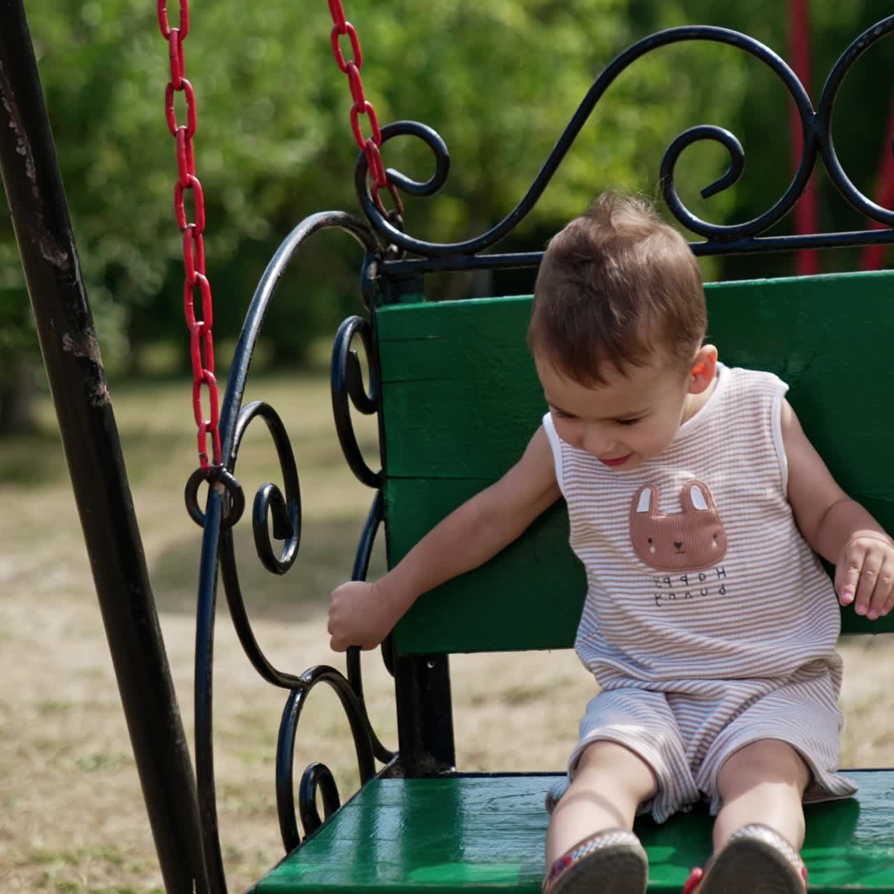 Adorable toddler boy swinging on the green swing in the park. Happy time outdoors in summer