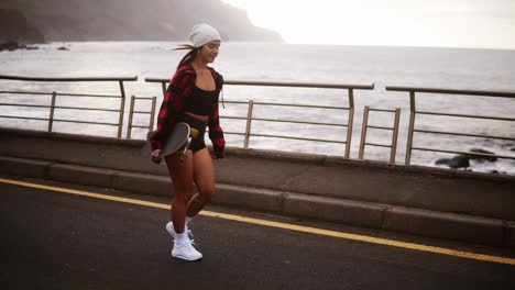 Mujer-Alegre-Y-Deportiva-Con-Sombrero,-Abrigo-Y-Pantalones-Cortos-Disfrutando-Del-Tiempo-Junto-Al-Mar-En-Un-Día-Nublado-Al-Atardecer,-Caminando-Libremente,-Sosteniendo-Una-Patineta