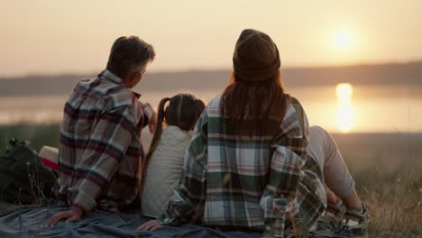 rear view of a happy family husband wife and their little daughter sitting on a mat during their vacation and picnic outside the city and looking at the sunset near a pond in the summer evening