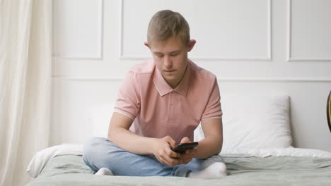 boy with down syndrome using smartphone sitting on the bed in the bedroom at home