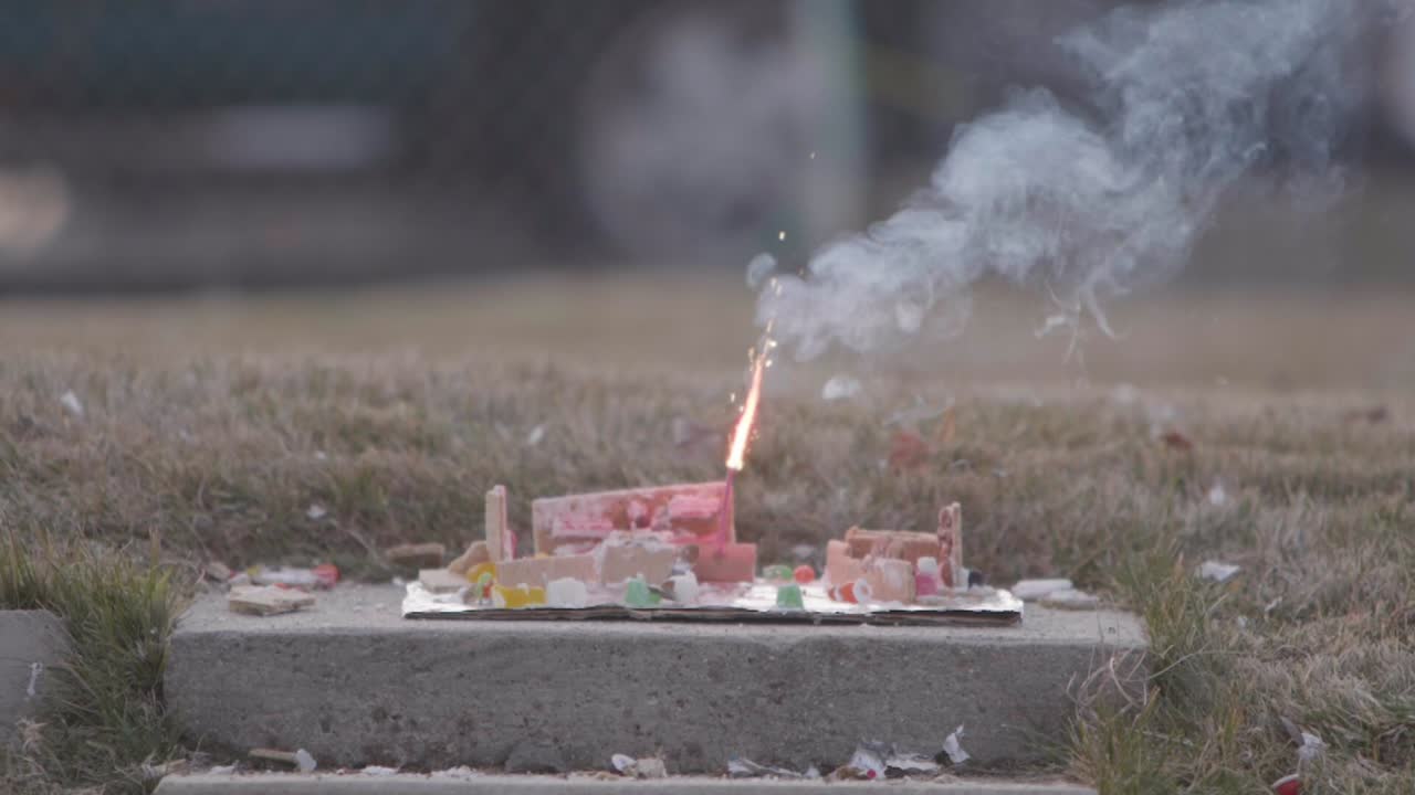 A Christmas ginger bread and graham cracker decoration has a large fire cracker or M 80 being lit on it by a flame from a blow torch
