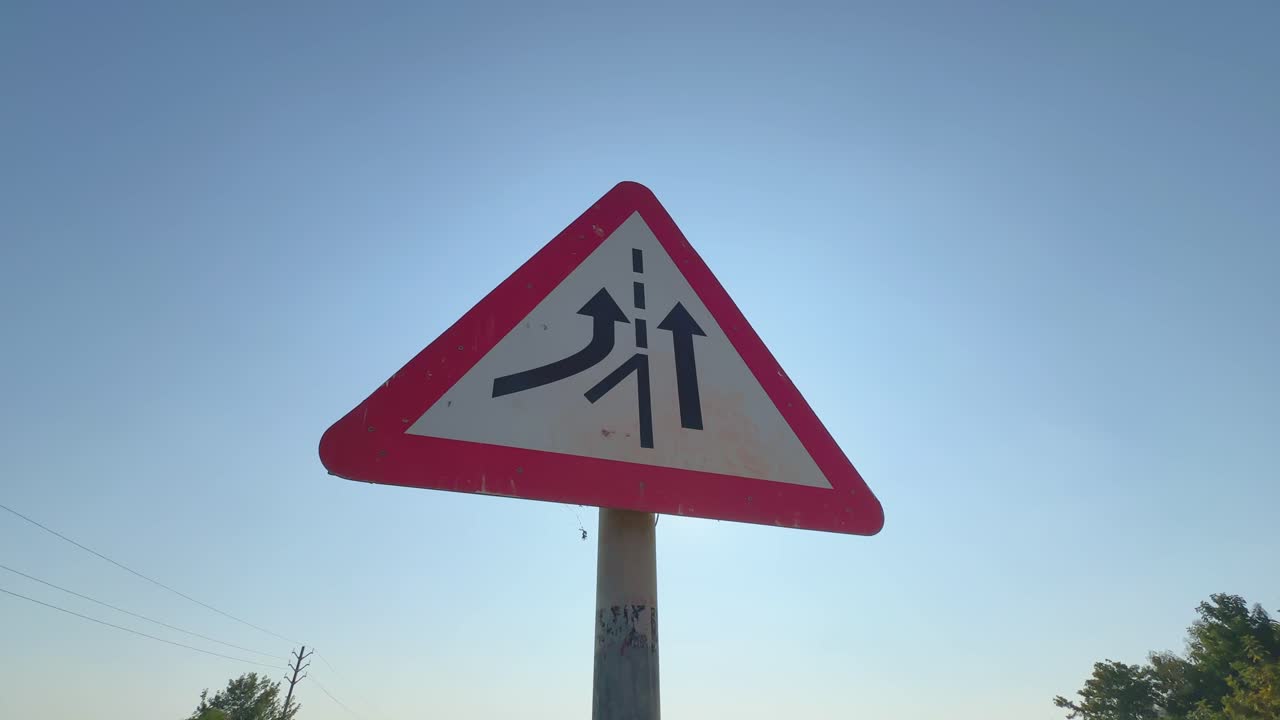 Low-angle orbiting shot around merging traffic ahead warning sign, black lane arrows clear against bright blue sky as camera circles smoothly emphasizing caution