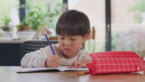 young asian boy home schooling working at table in kitchen writing in book during lockdown