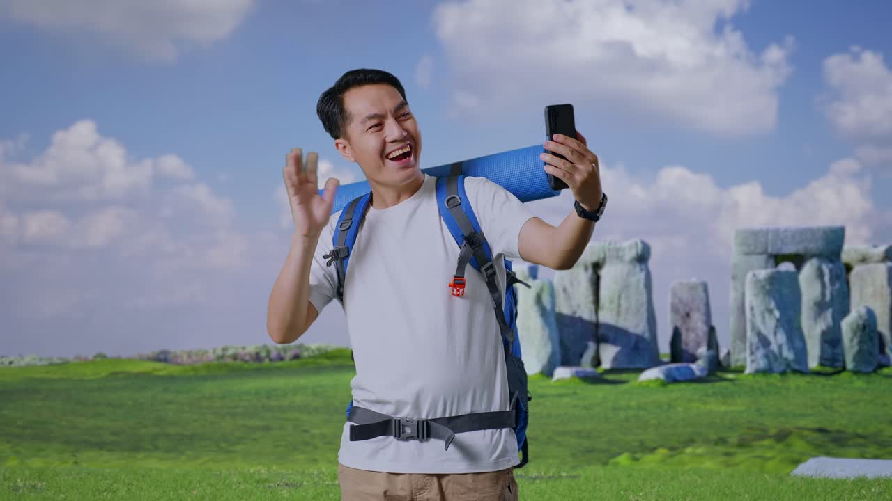 Asian Male Hiker With Mountaineering Backpack Waving His Hand And Pointing Back While Having A Video Call On Smartphone While Traveling In Stonehenge
