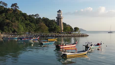 Hermosa-Vista-Aérea-De-La-Puesta-De-Sol-Del-Faro-Del-Muelle-Perdana-Con-Barcos-De-Pesca-Tradicionales-Amarrados-A-La-Orilla-Con-Dos-Veleros-Navegando-Desde-El-Puerto-Deportivo-De-Telaga-En-La-Isla-Langkawi,-Kedah,-Malasia