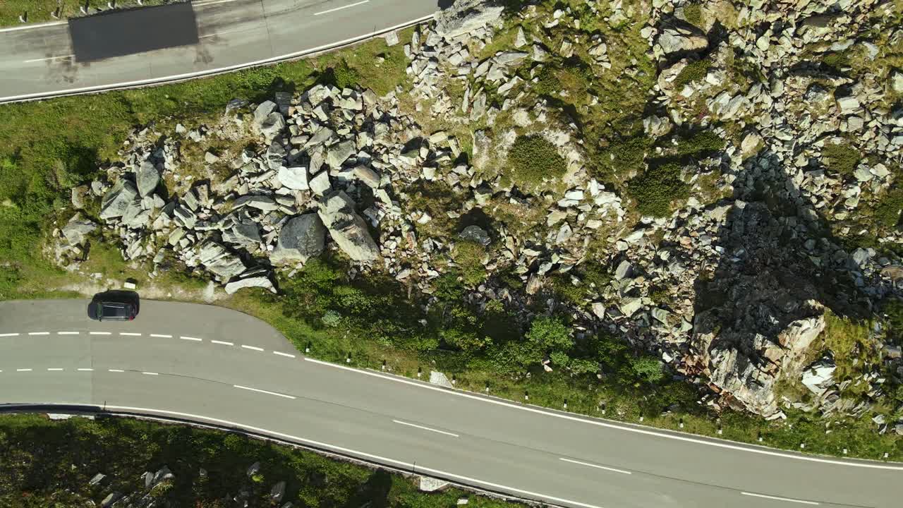 A top-down aerial view of a tight switchback curve on a mountain road, with several cars navigating the bend. The rocky terrain and sparse vegetation highlight the ruggedness of the alpine environment