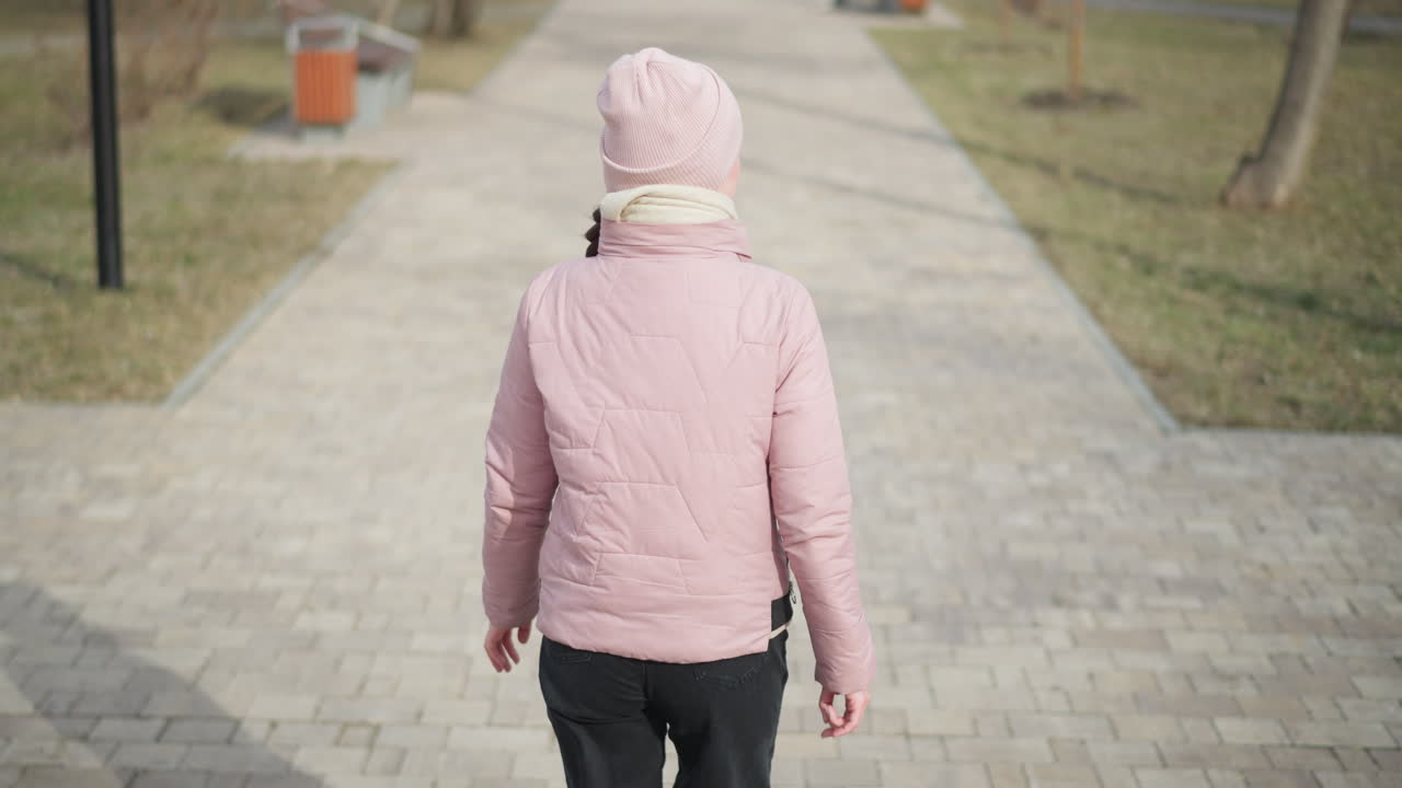 Woman in pink jacket and beanie walks away on empty paved park path during chilly winter day, surrounded by bare trees and soft sunlight, representing solitude, calmness, outdoor lifestyle, and seasonal fashion