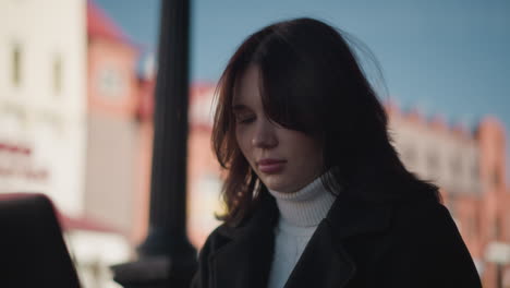 close-up of a woman with dark hair and a thoughtful expression sitting outdoors in soft light, she is dressed in a black coat and white turtleneck, with a blurred urban background