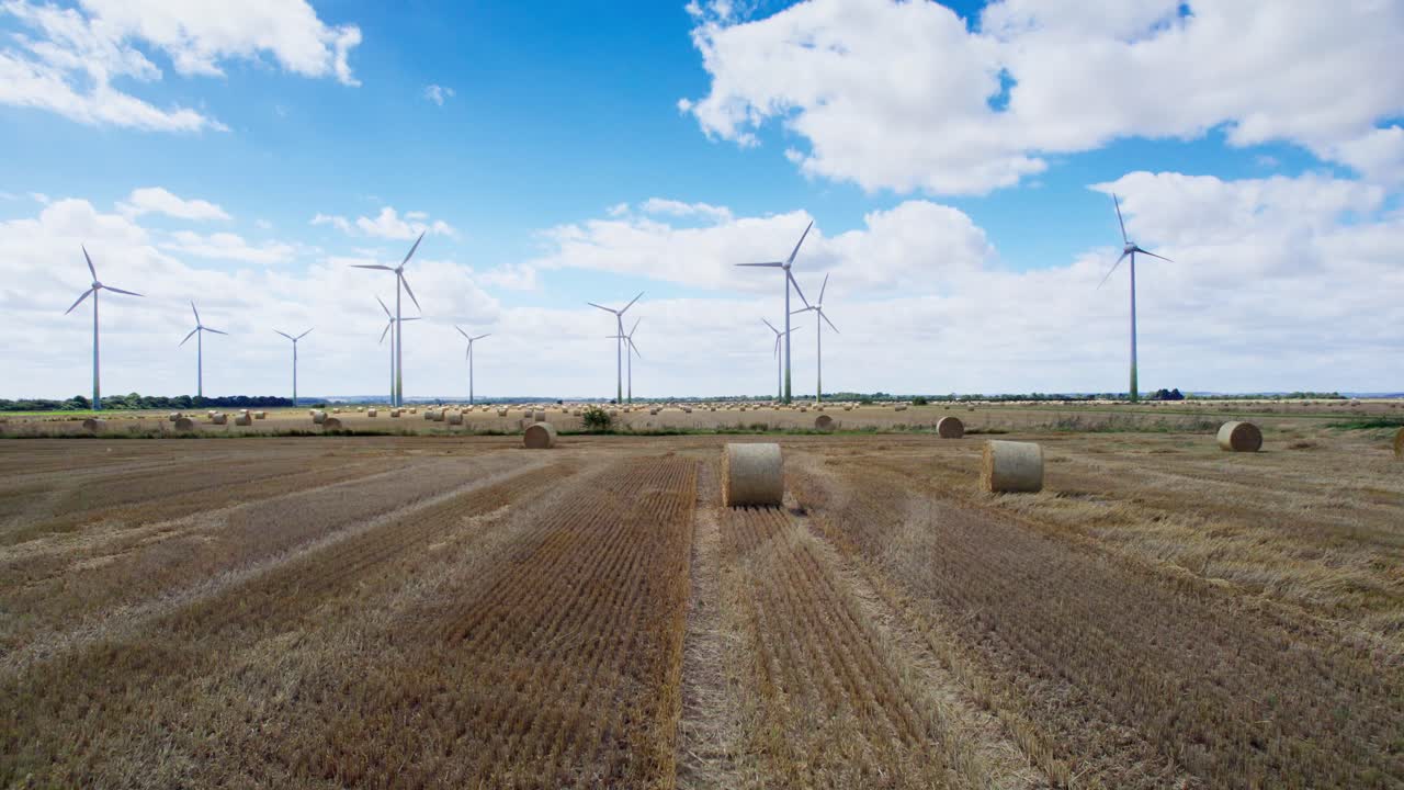 In this aerial video, wind turbines stand tall and spin in a Lincolnshire farmer's newly harvested field, framed by golden hay bales in the foreground