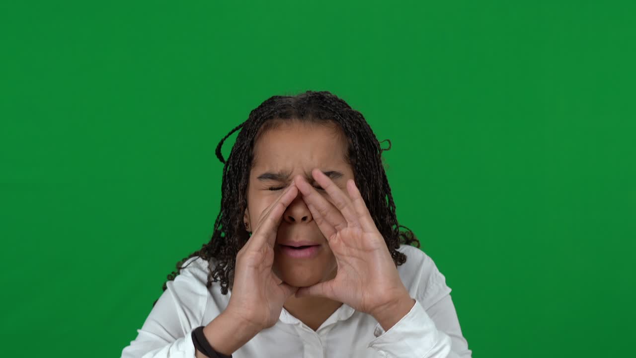 Close-up of stressed lost African American teen girl shouting calling for help at chromakey background. Upset sad disoriented teenager on green screen yelling holding hands at mouth.