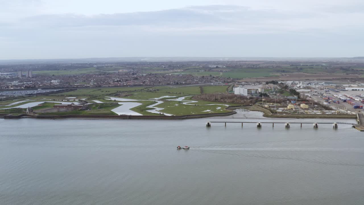 Drone slow dolly over boat on calm River Thames near Gravesend showing green riverbanks and docks