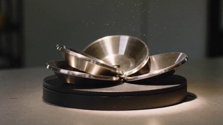 Hinge releasing, three-steel petal assembly unfolding on black plinth, showing dust and reflections