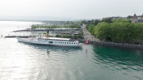 Drone-flying-away-from-a-beautiful-cruise-ship-docked-at-harbor-and-revealing-the-city-skyline-of-Lausanne,-Switzerland-at-lake-Geneva