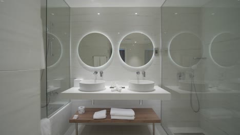 a interior gimbal shot of a beautiful elegant glassy hotel bathroom full of reflections inside of a resort apartment with two round mirrors and white porcelain sinks