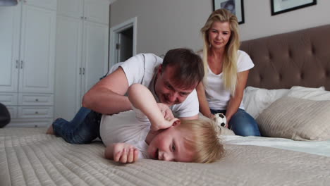 mom and father tickling her child. people, family, fun and morning concept - happy child with parents tickling in bed at home. happy family spending time together in bedtime playing and hugging