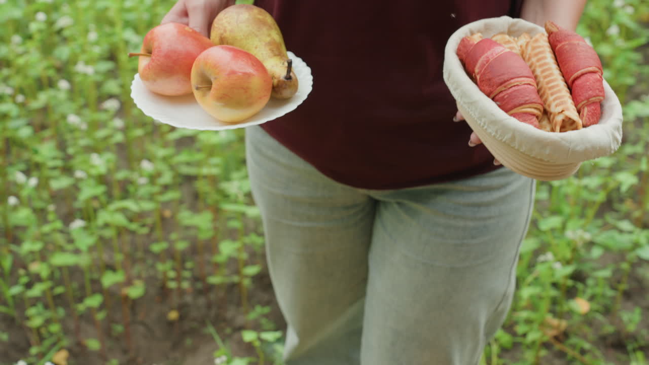 Mujer caucásica organizando fruta para un picnic, arrodillada para colocar una cesta y un plato sobre una toalla con una sandía cerca, con vaqueros y sandalias informales, las manos ajustando manzanas y cucuruchos, ambiente acogedor al aire libre.