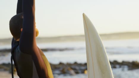 Vista-Lateral-De-Un-Surfista-Caucásico-De-Mediana-Edad-Estirándose-Y-Calentándose-Antes-De-Surfear-En-La-Playa-4k