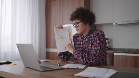 A-curly-man-with-glasses-at-his-desk-shows-a-picture-with-graphs-in-a-laptop-camera