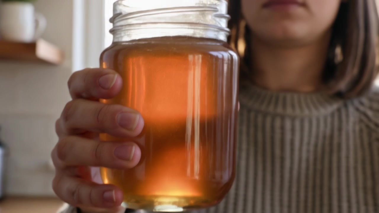 Donna che si gira con un top a costine, mentre svita il coperchio da un barattolo di vetro in cucina, ispezionando un liquido ambrato.