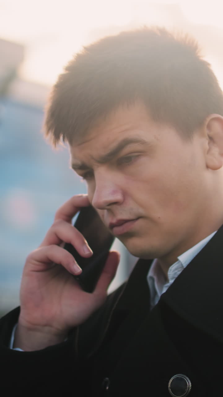 검은 코트를 입은 투자자는 전화를 할 때 머리를 끄덕이는 동안  아래에 전화를 들고 동의하는 것을 나타니다. 배경에는 현대적인 유리 건물과 부드러운 빛이 있습니다.