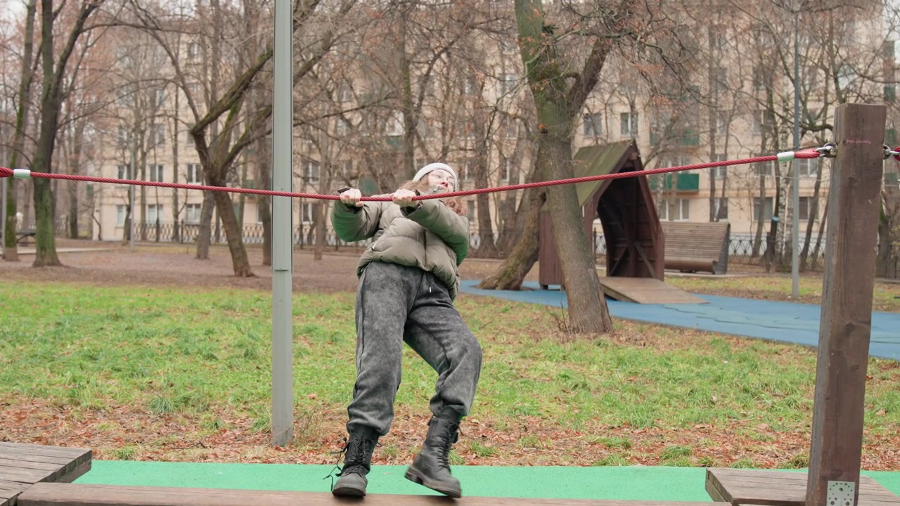 White woman balancing on red rope, winter playground practice bundled in puffy jacket and knit hat, carefully stepping across low rail while gripping rope for stability, candid sequence shows playful