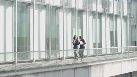 wide shot of two businesswomen walking together on office building glass terrace, holding papers, tablet and talking