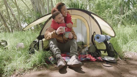 una feliz pareja afroamericana acampando, bebiendo café y abrazándose en el bosque, en cámara lenta.