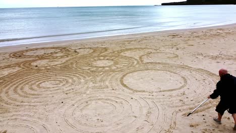 Luftaufnahme-Eines-Umkreisenden-Mannes,-Der-Am-Windigen-Walisischen-Goldenen-Anglesey-Strand-Ein-Interessantes-Zen-Sand-Kunstdesign-Kreiert