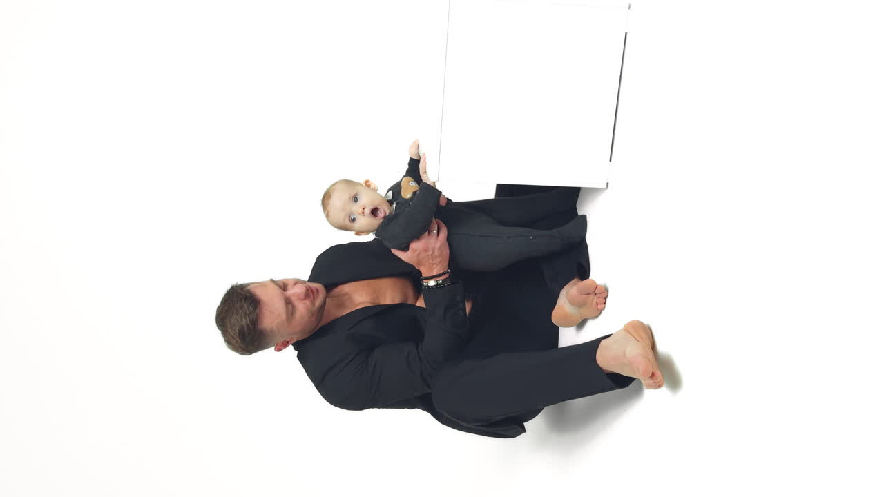 Caucasian man in black suit sits on the floor with a tiny baby. Little infant holds by the white cube. White backdrop. Vertical screen.